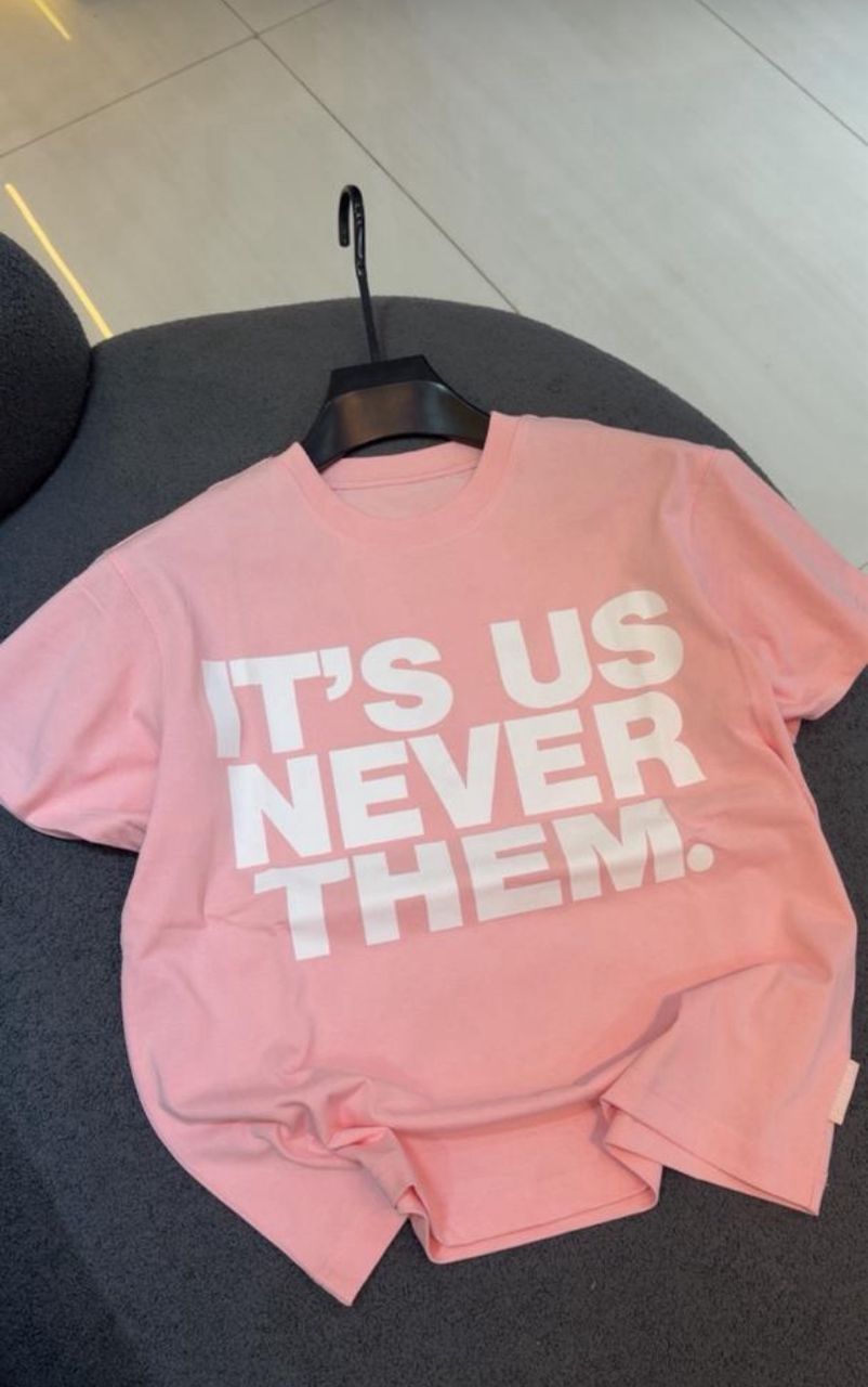 Pink sweatshirt with 'IT'S US NEVER THEM' text on a hanger against a neutral background