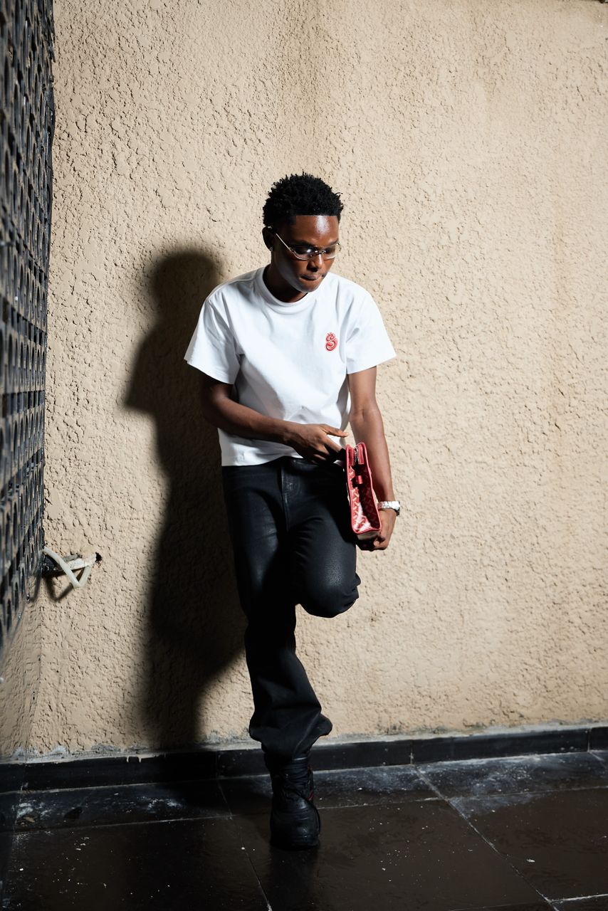 SBM wearing a white t-shirt and black pants against a textured wall.