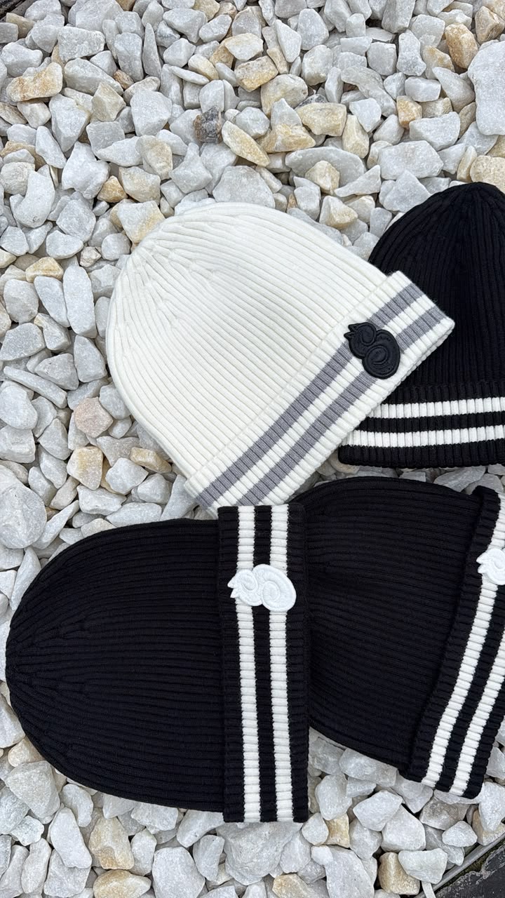 White knit beanie with gray stripes and black pom-pom, paired with black sandals on a pebbled surface.